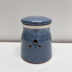 May include: A blue ceramic garlic keeper with a matching lid. The keeper has three small holes and a white band near the top. The ceramic has a speckled texture and a glossy finish. The keeper is sitting on a white surface.