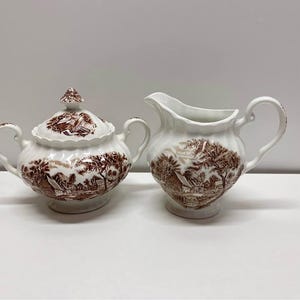 May include: A white ceramic sugar bowl with a lid and a creamer. Both items display a brown transferware rural landscape design. The sugar bowl has two handles and a decorative finial. The creamer has a handle and a pouring spout.