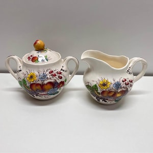 Peut inclure: Un sucrier et un pot à crème vintage de couleur crème. Les deux pièces sont décorées d'un motif floral et fruité coloré, comprenant des fleurs jaunes, des baies rouges et des pêches. Le sucrier a deux anses et un petit bouton sur le couvercle.