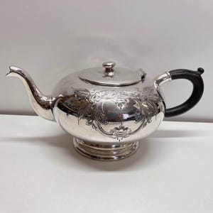 Op de afbeelding: Een zilveren theepot met een zwarte handgreep en een bloemmotief. De theepot heeft een deksel en een tuit. De theepot staat op een wit oppervlak.