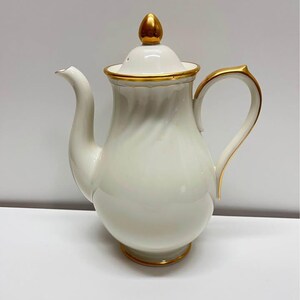May include: A white porcelain coffee pot with gold trim and a gold finial on the lid. The handle and spout are also trimmed in gold. The pot has a bulbous body and a curved spout, suitable for serving coffee or tea.