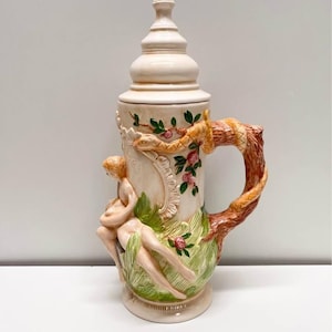 Może przedstawiać: Kremowy ceramiczny kufel do piwa z płaskorzeźbą przedstawiającą dwie postacie leżące w zielonej trawie. Uchwyt ma kształt teksturowanej brązowej gałęzi drzewa owiniętej wężem. Pokrywka to pasujące kremowe, spiczaste zwieńczenie.