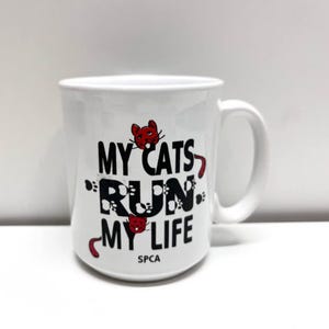 May include: A white ceramic mug featuring the black text "MY CATS RUN MY LIFE" with two small red cat illustrations and black paw prints integrated into the lettering. The acronym "SPCA" is printed in small black letters at the bottom center.
