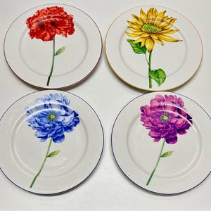 May include: A set of four round white ceramic plates, each featuring a single botanical illustration of a flower. The top left plate displays a red poppy, the top right a yellow sunflower, the bottom left a blue peony, and the bottom right a purple peony.