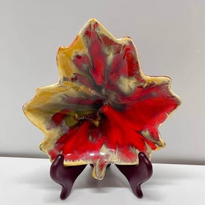 May include: A leaf-shaped ceramic dish featuring a glossy glaze with blended colours of vibrant red, golden yellow, and dark brown. The dish has an organic, wavy edge and is displayed on a dark purple plastic stand, showcasing its unique form.