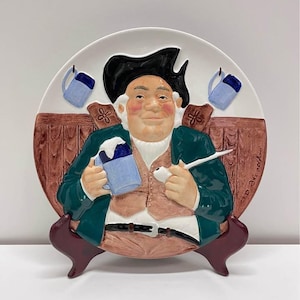 Puede incluir: Plato decorativo de cerámica que representa a un hombre jovial con un abrigo verde, sosteniendo una jarra azul y una pipa. El plato tiene un fondo de madera marrón y dos pequeñas tazas azules colgando en la parte superior. Soporte burdeos.