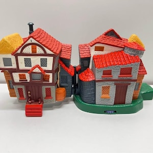 May include: A whimsical miniature village scene featuring two connected buildings. The buildings have red tile roofs, tan and gray walls, and small windows. One building has a yellow thatched roof. The scene is set on a green base.