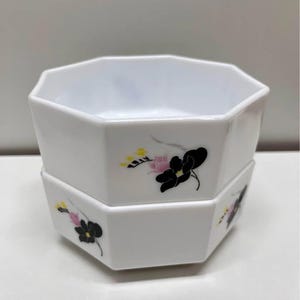 May include: Two white, octagonal bowls stacked on top of each other. Each bowl is decorated with a black floral design with pink and yellow accents. The bowls have a glossy finish and are set against a plain background.