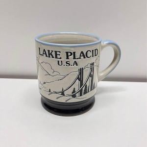 May include: A ceramic mug with a light beige body, a black base, and a light blue rim and handle. The mug features a black and white graphic of a bridge and mountains, with the text "LAKE PLACID U.S.A."