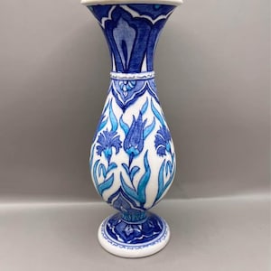 Puede incluir: Un jarrón de cerámica blanca con la parte superior acampanada y una base redonda. El jarrón está adornado con un patrón floral azul y turquesa. El diseño incluye flores y hojas estilizadas, con una banda azul en el cuello y la base.