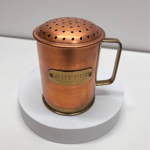 May include: A copper cheese shaker with a perforated lid and a handle. The word "CHEESE" is displayed on a brass plate. The shaker is made of copper and brass.