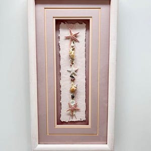 May include: A framed artwork with a coastal theme, showcasing a vertical display of seashells and starfish. The piece features three red starfish and assorted seashells on a textured, pale pink backdrop. The frame is light pink with a white outer frame.