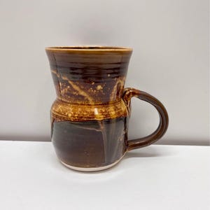 May include: A ceramic mug with a glossy finish featuring a two-tone brown glaze. The upper section has a lighter, mottled tan and brown pattern, while the lower section is a solid, darker chocolate brown with visible drip-like glaze effects.