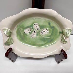 May include: An oval ceramic dish with a cream-coloured rim and a light green glazed centre. The centre features a hand-painted stem with four white, leaf-shaped accents. Two small, light green, wavy-textured leaves are attached to each side.