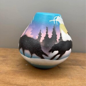 Può includere: Un vaso in ceramica dipinto a mano con un design paesaggistico. Il vaso presenta un gradiente di tonalità blu, viola e rosa, con sagome nere di alberi e animali. La base è azzurra e bianca.