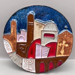 May include: A round, colorful ceramic tile featuring an abstract depiction of a cityscape. The design includes buildings, a cross, and a menorah, with the name "YAIR" on the left. The tile has a blue, brown, red, and white color scheme.
