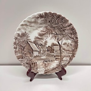 May include: A brown and white decorative plate featuring a rural landscape scene with a watermill, trees, and a waterfall. The plate has a scalloped edge and is displayed on a dark red stand. The plate's diameter is approximately 10 inches.