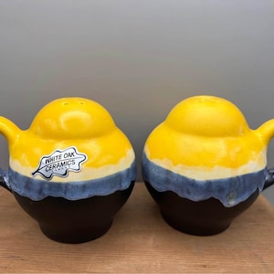 May include: Two ceramic salt and pepper shakers. Each shaker features a yellow top, a blue band, and a dark brown base. The shakers have handles and the text "WHITE OAK CERAMICS" is visible on one.