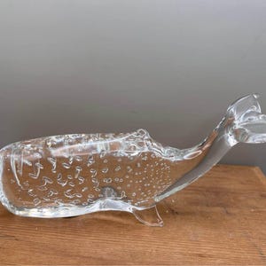 May include: A clear glass whale-shaped decorative bowl. The whale has a curved body, a tail, and a small fin. The glass has bubbles throughout. The bowl is sitting on a wooden surface.