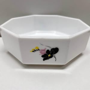 May include: White, octagonal serving bowl with a floral design. The bowl has a simple, geometric shape and is decorated with a black, pink, and yellow flower on the side. The bowl is likely made of ceramic or porcelain.