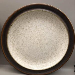 May include: A round ceramic plate with a speckled off-white center and a dark brown rim. The plate has a wide, dark brown border and a slightly raised edge. This plate is suitable for serving meals.
