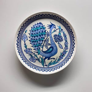May include: A round, white ceramic plate with a hand-painted peacock design. The peacock is depicted in shades of blue and teal, surrounded by floral elements and a decorative border. The plate is a decorative item.