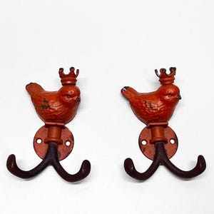 May include: A pair of wall hooks made of cast metal, each featuring a bird figure wearing a small crown on its head. The birds are painted in a distressed orange finish, while the double-pronged hooks below are a dark brown colour. Each hook has a circular mounting plate with two screw holes.
