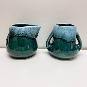 May include: A ceramic creamer pitcher and a sugar bowl set featuring a deep emerald green base glaze that transitions into a lighter, pale blue drip pattern at the top. The creamer has a pouring spout, while the sugar bowl has two side handles.