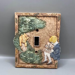 May include: Decorative light switch cover with a relief design of a bear and a child reading. The cover is rectangular with a textured, wood-like finish. The bear is yellow, and the child is wearing blue. Green foliage forms the background.