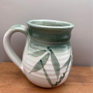 Peut inclure: Mug en céramique fait à la main avec une base blanche et des accents verts. La tasse présente un bord et une anse verts, avec un motif de feuilles peintes dans des tons de vert. La tasse mesure environ 10 cm de haut et 8 cm de diamètre.