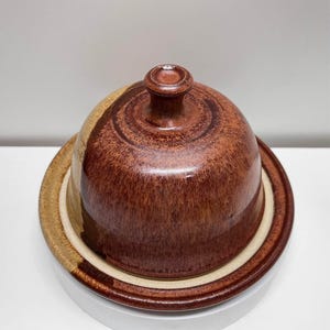 May include: A round ceramic butter dish with a dome-shaped lid featuring a knob handle. The piece has a mottled glaze finish in shades of deep reddish-brown and warm tan, with a smooth, glossy texture covering the entire surface of the lid and base.