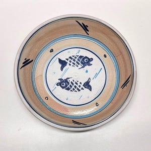 May include: A round ceramic bowl featuring a light tan interior with a central white circle. Two dark blue fish motifs are painted in the center, surrounded by light blue rings and small dark blue dash accents along the rim of the bowl.