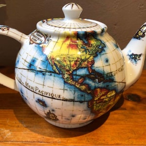 May include: A white ceramic teapot with a globe design. The teapot features a handle, spout, and lid, all adorned with a world map in shades of blue, yellow, and green. The words "MER PACIFIQUE" are visible on the teapot.