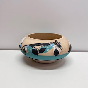 May include: A beige ceramic bowl with a turquoise band and feather design. The bowl features black and white feathers with blue accents. The bowl is round with a wide opening and sits on a white surface.