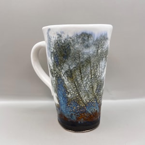May include: Tall white ceramic mug with a unique design. The mug features a white handle and a body adorned with a textured pattern in shades of blue, green, and brown. The rim is white, and the base is dark brown.