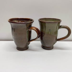 May include: Two handmade ceramic mugs with a brown and green glaze. The mugs have rounded bodies, curved handles, and a slightly flared rim. The mugs are displayed on a white surface.
