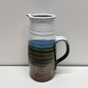 May include: A tall ceramic pitcher featuring a cylindrical body with horizontal ridges. The glaze transitions from a white top to a blue middle section, ending in a dark brown and grey base. It includes a curved handle and a small pouring spout.