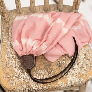 May include: A pink and white tie-dye scarf with a brown leather camera strap, draped over a distressed wooden chair. The scarf has a soft, flowing texture and a pattern of white cloud-like shapes.