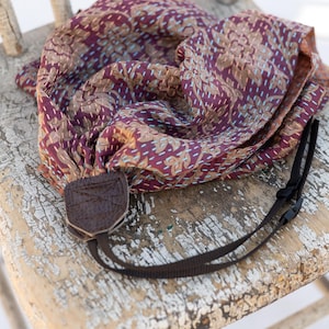 Pode incluir: Uma alça de câmera estampada com um conector de couro marrom e uma alça de nylon preta repousa sobre uma cadeira de madeira desgastada. O tecido é uma mistura de bordô, bege e branco, com um design floral e costuras visíveis.