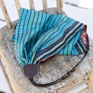 Pode incluir: Um lenço azul e turquesa estampado com uma alça de câmera de couro marrom está sobre uma cadeira de madeira desgastada. O lenço apresenta um design costurado e um interior estampado contrastante.