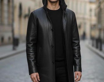 May include: A black leather hooded coat with a button-up front. The coat is long, reaching the mid-thigh, and is worn over a black shirt and pants. The person is standing on a cobblestone street.