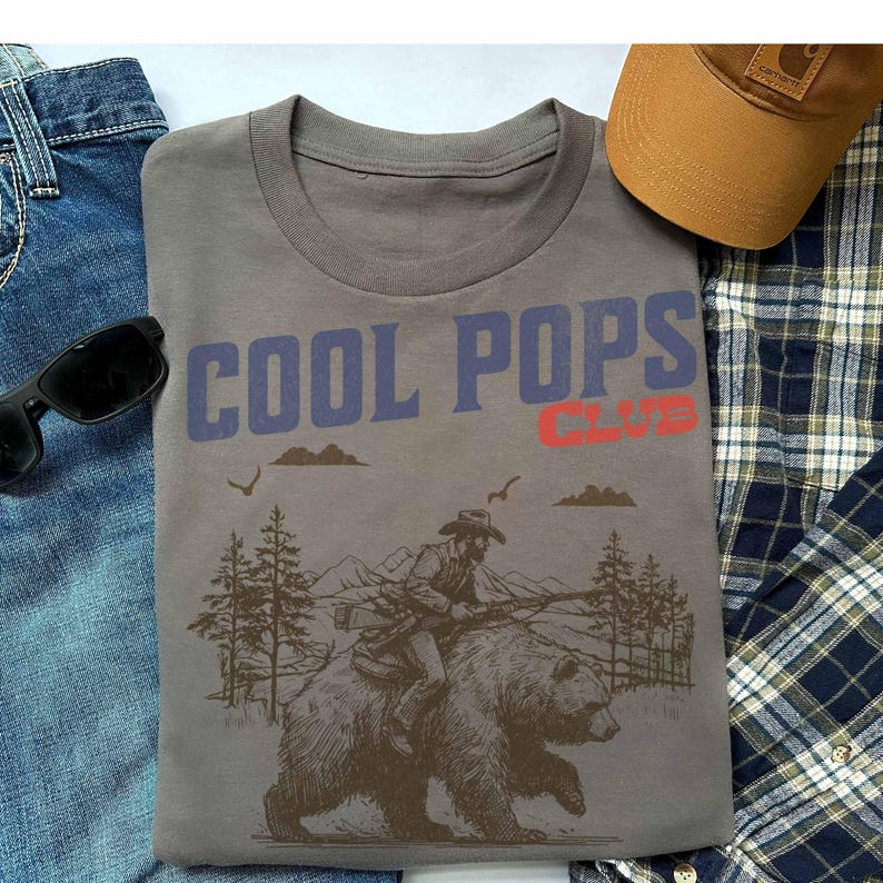 May include: A gray t-shirt with a graphic of a cowboy riding a bear. The text "COOL POPS CLUB" is printed in blue and red on the shirt.
