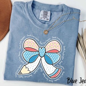 May include: Blue jean coloured t-shirt featuring a colourful bow and pencil graphic. The bow has pink, blue, and red accents with a polka dot pattern. The words "Teach Love Inspire" are printed around the bow. A gold necklace is draped over the shirt.