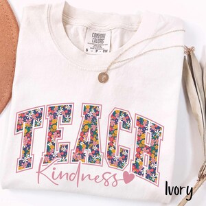 May include: Ivory-coloured t-shirt featuring the word "TEACH" in a floral design, and the word "Kindness" in a cursive font. A small pink heart is below "Kindness". A gold necklace is draped on the shirt.