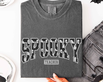 Puede incluir: Una camiseta de cuello redondo gris carbón con la palabra SPOOKY en letras grandes de bloque rellenas con un patrón de fantasmas blancos. Debajo de la palabra SPOOKY, la palabra TEACHER está impresa en un pequeño recuadro blanco.