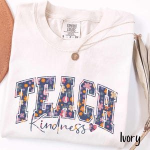 May include: An ivory-coloured t-shirt featuring the words "TEACH Kindness" in a floral design. The word "TEACH" is in a navy blue, collegiate-style font, and "Kindness" is in a script font. A gold necklace is draped across the shirt.