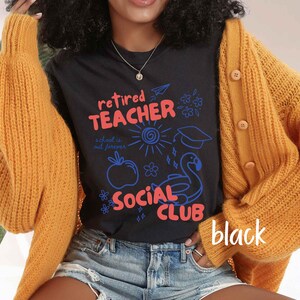 May include: Black t-shirt with the words "Retired Teacher Social Club" in red and blue, with illustrations of a sun, apple, graduation cap, and flamingo. The shirt is worn with a gold cardigan and denim shorts.