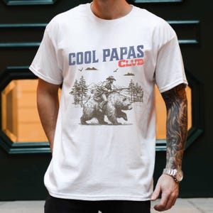 May include: White t-shirt featuring a graphic of a person riding a bear through a forest. The text "COOL PAPAS CLUB" is printed on the shirt.