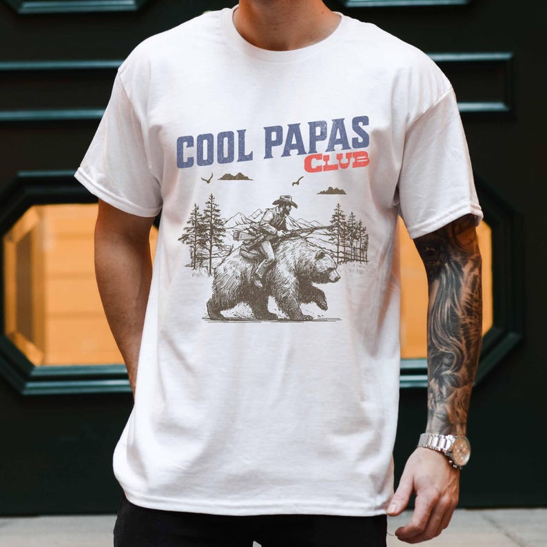 May include: White t-shirt featuring a graphic of a person riding a bear through a forest. The text "COOL PAPAS CLUB" is printed on the shirt.