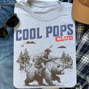May include: Gray t-shirt with a vintage-style graphic of a cowboy riding a bear. The text "COOL POPS CLUB" is printed in blue and red on the shirt.